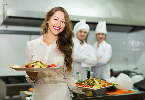 Positive waitress take plates with prepared meal at kitchen